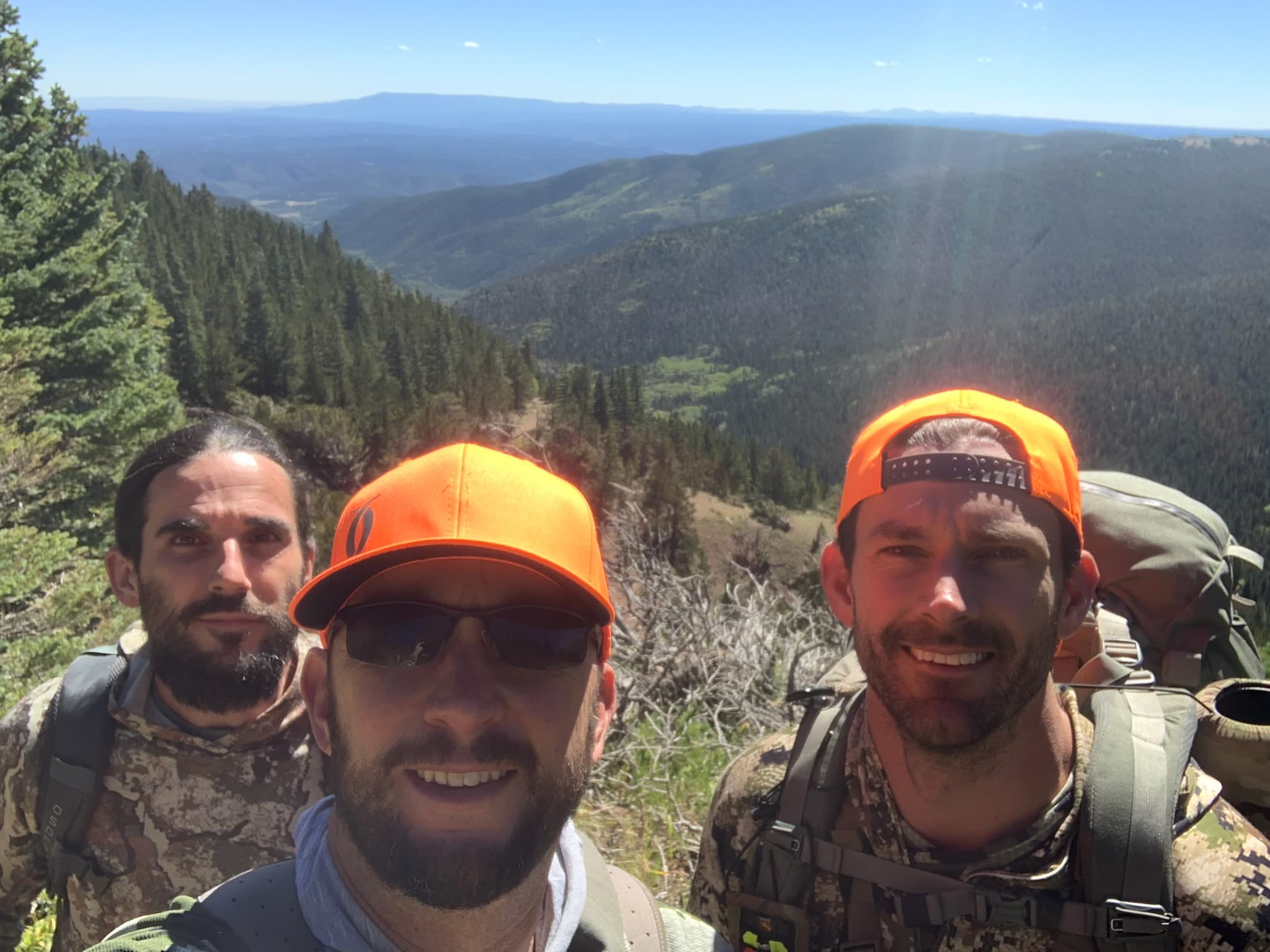 Three backcountry hunters in blaze orange caps and packs on an alpine ridge, mountains stretching to the horizon