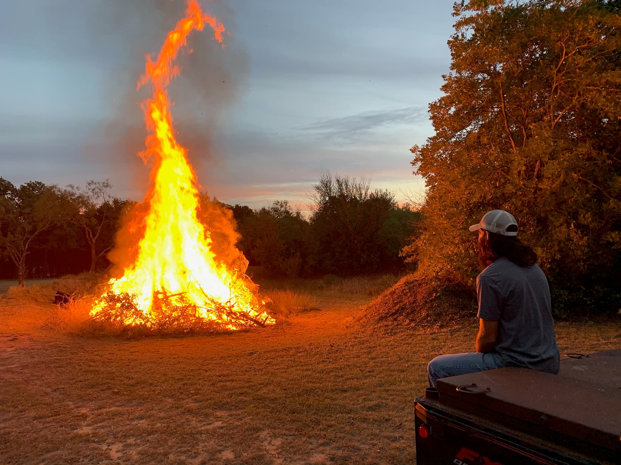 Man on a tailgate watching a brushpile bonfire at dusk
