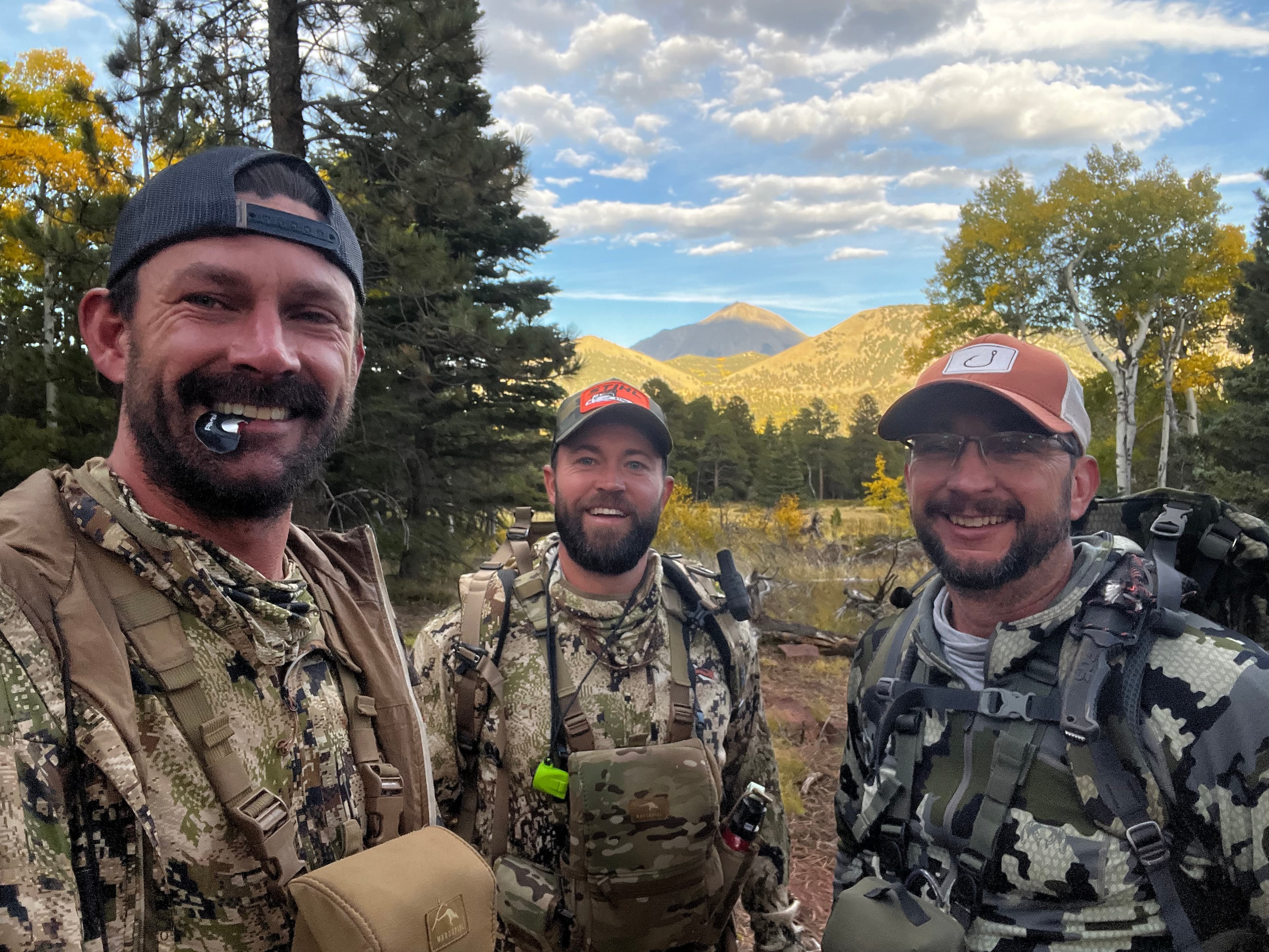 Three men in camo gear with blue sky and aspens