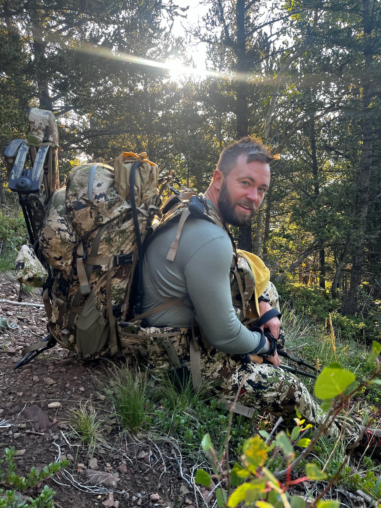 Bowhunter with a full pack glancing back as sun flares through pines