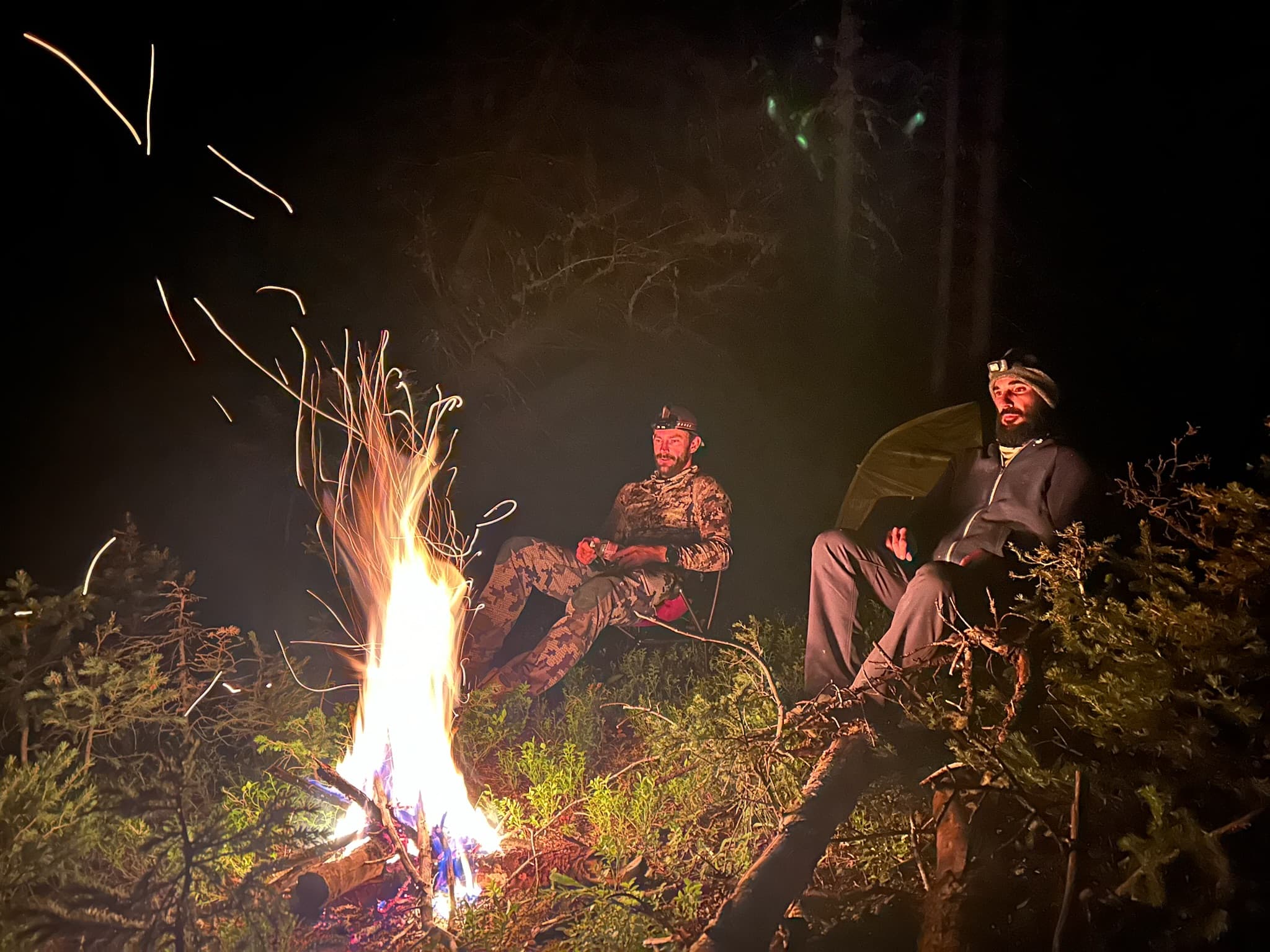 Two practitioners around a campfire at night, sparks rising into the trees