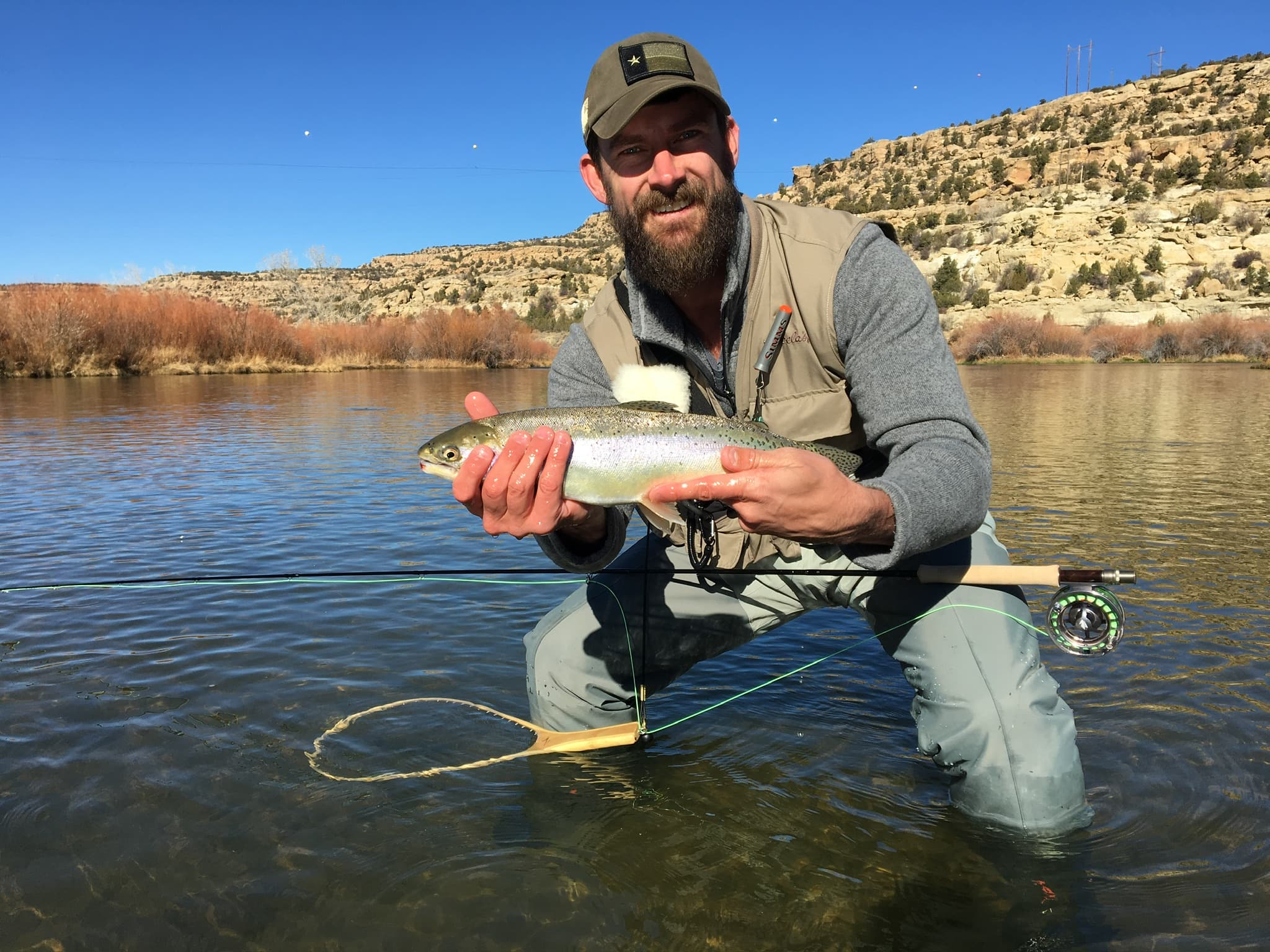 Fly angler kneeling in a river with a rainbow trout, red rocks