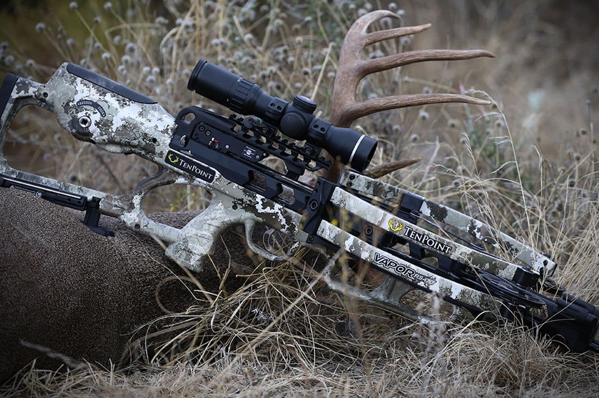 A camouflaged TenPoint Vapor crossbow at rest beside a set of antlers in tall winter grass