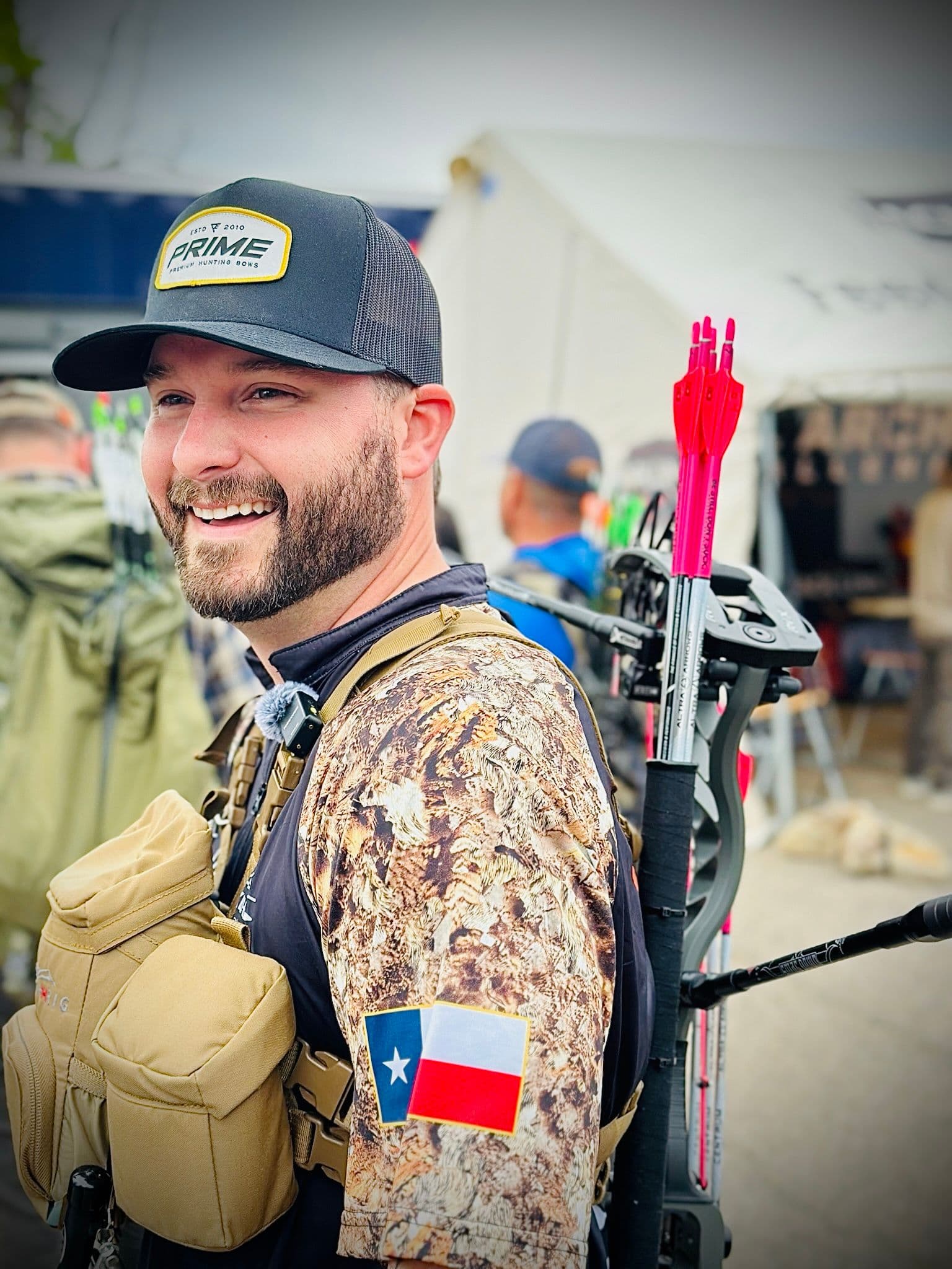 Justin, owner of Bear River Archery, in a Prime hat with a compound bow and pink-fletched arrows over his shoulder, wearing a Texas flag patch