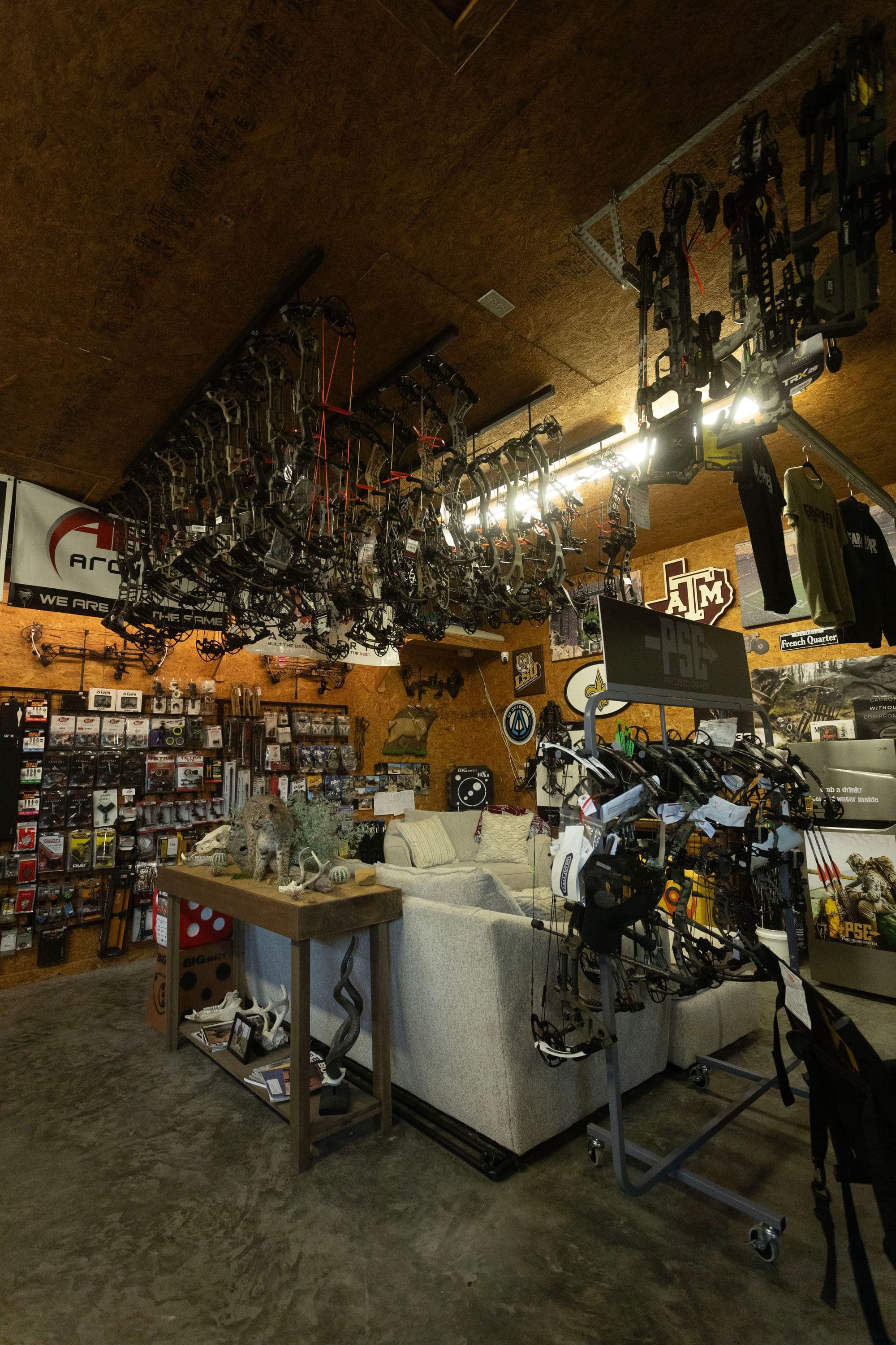Inside Bear River Archery: compound bows hanging from the ceiling rail, gear walls, a Texas A&M flag, accessory pegboards, and a workbench in the center of the shop